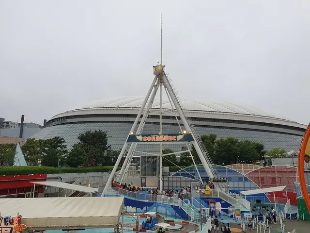 The low white roof of Tokyo Dome behind a tall A-frame swing ride and other fairground rides under a grey sky