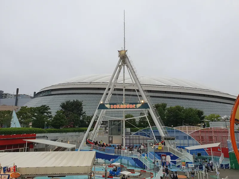 The low white roof of Tokyo Dome behind a tall A-frame swing ride and other fairground rides under a grey sky