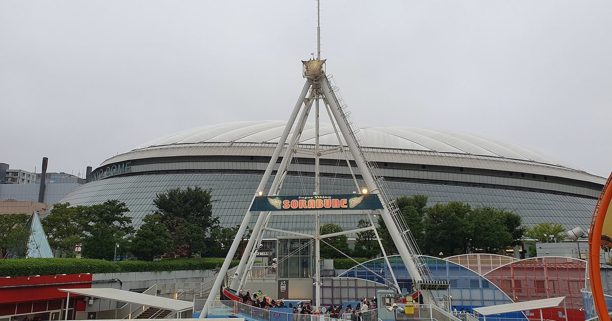 The low white roof of Tokyo Dome behind a tall A-frame swing ride and other fairground rides under a grey sky