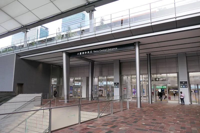 A wide station entrance under a white canopy, with steel columns, glass doors, a brick-paved forecourt and a sign reading Yaesu Central Entrance
