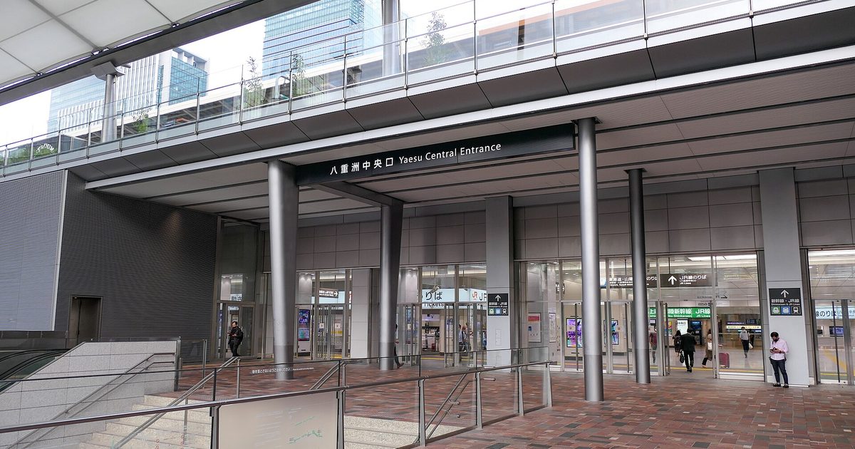 A wide station entrance under a white canopy, with steel columns, glass doors, a brick-paved forecourt and a sign reading Yaesu Central Entrance