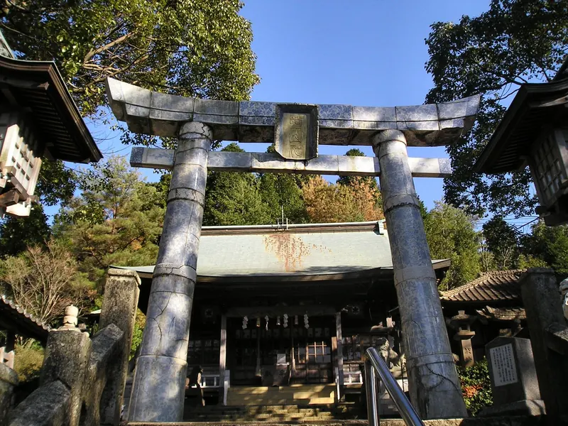 A pale grey torii gate built of stacked sections in front of a shrine hall among trees
