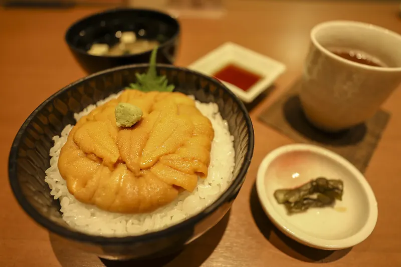 A black bowl of white rice covered with orange sea urchin and a dab of wasabi, with soup, pickles, a dipping sauce and a cup of tea beside it