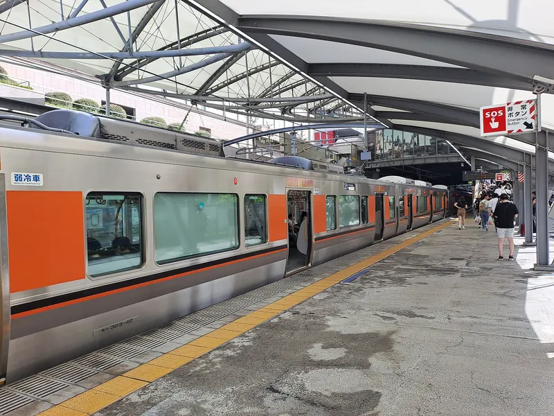 A silver JR commuter train with orange panels at an open-air platform under a white canopy roof