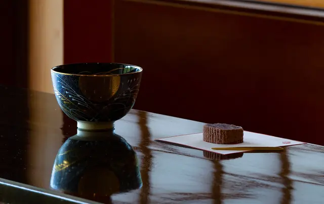 A dark tea bowl and one sweet on paper on a glossy lacquer table