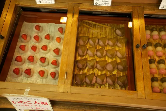 A wooden display case with trays of strawberry daifuku, pink rice cakes wrapped in leaves and three-colour dango
