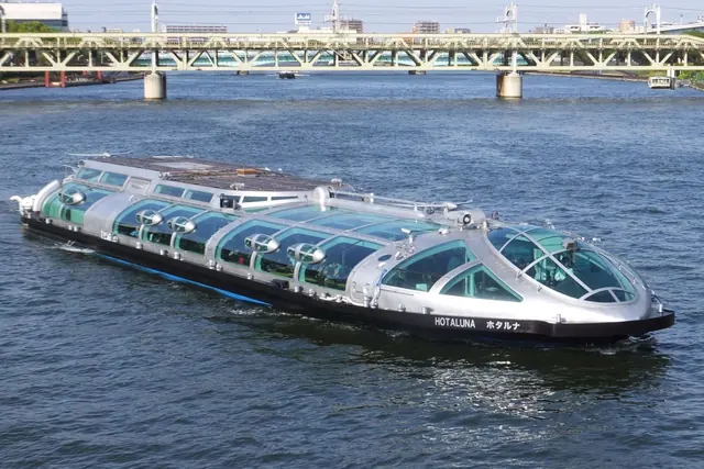 A low silver boat with a glass roof and the name Hotaluna on its hull, on a river with a green bridge behind