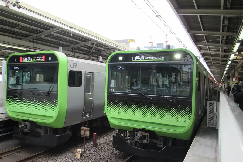 Two light-green and silver commuter trains side by side at a covered platform, one with Yamanote Line on its front display