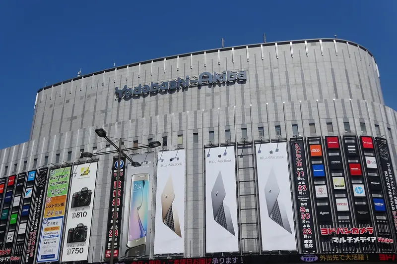 A wide grey curved building with the name Yodobashi-Akiba near the roof and rows of tall advertising banners along its lower floors, under a blue sky
