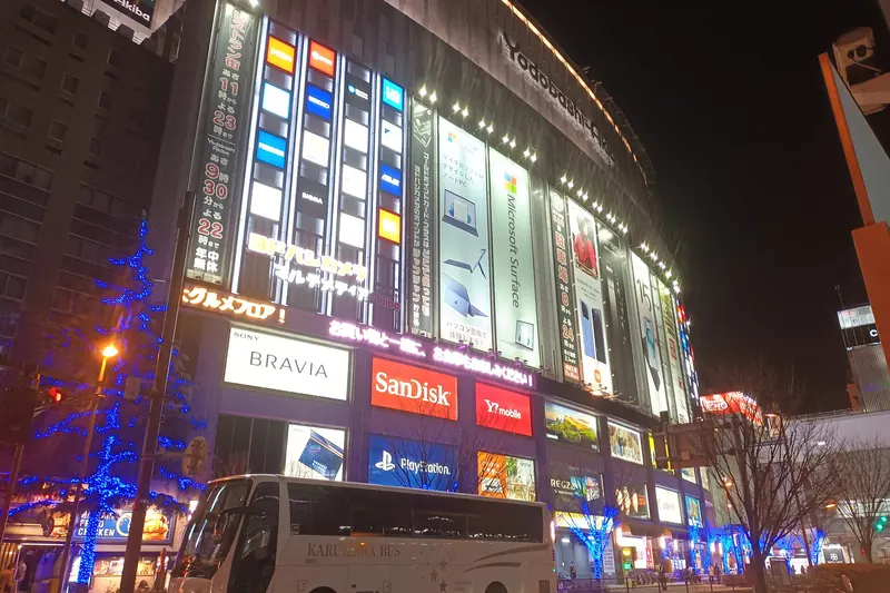 A large electronics store at night, its front covered in lit advertising panels, with a white coach parked in front and blue lights on the street trees