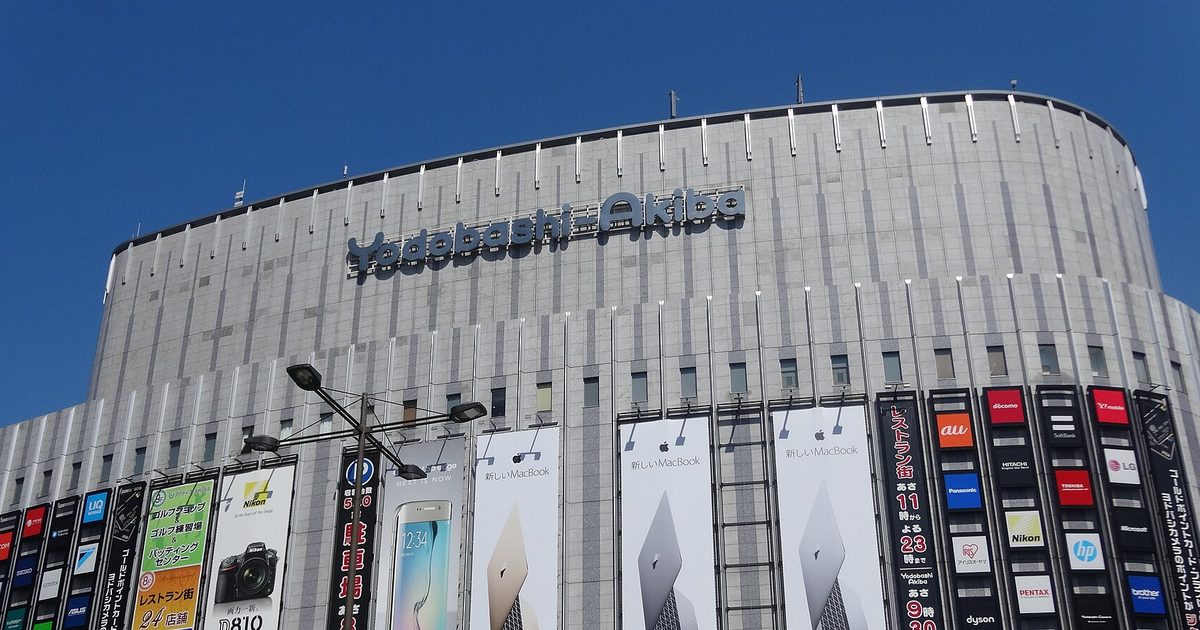 A wide grey curved building with the name Yodobashi-Akiba near the roof and rows of tall advertising banners along its lower floors, under a blue sky