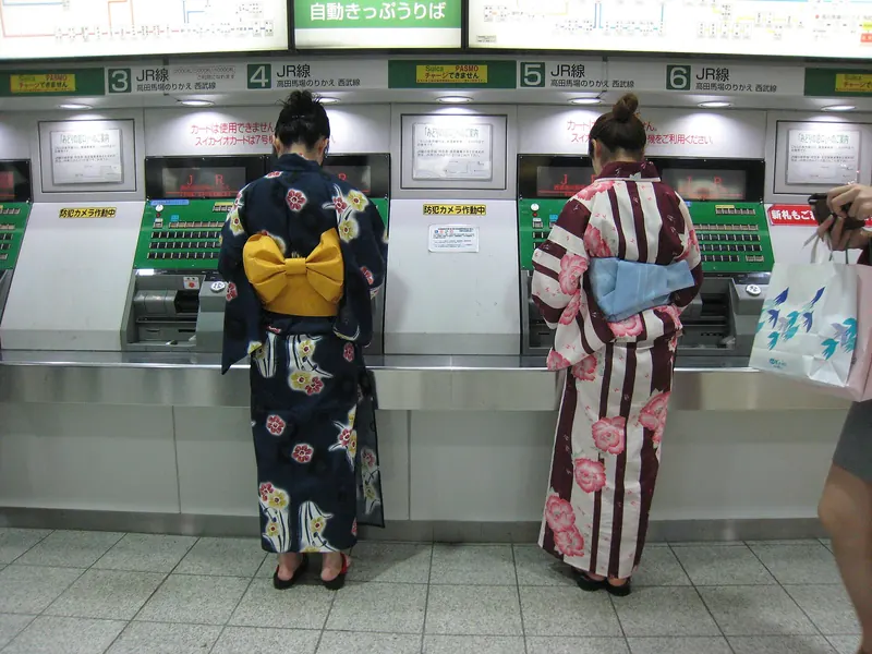 Two women in patterned yukata with obi seen from behind at ticket machines